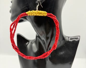May include: A large hoop earring featuring a textured red twisted cord frame. The top section of the hoop is wrapped in a metallic gold thread. The earring is attached to a silver-toned metal hook for pierced ears, showcasing a bold design.