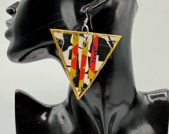 May include: A triangular dangle earring featuring a collage of black, white, red, and yellow paper elements with visible text fragments including the word SHAKES. The earring has a gold-toned border and hangs from a silver-toned metal hook.