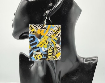 May include: A square-shaped dangle earring featuring a collage of printed paper with yellow, blue, black, and white text and graphics. The earring hangs from a silver-toned metal hook and displays fragments of words including SOCIAL, TRUCK, and OLLIE'S.