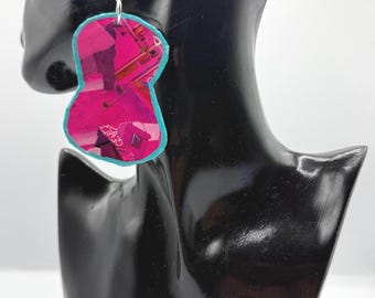 May include: A dangling earring featuring an irregular, hourglass-shaped pendant with a vibrant magenta and dark purple collage pattern. The pendant is outlined with a bright turquoise blue border and hangs from a silver-toned metal hook wire.