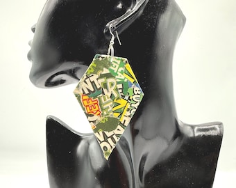 May include: A diamond-shaped dangle earring featuring a collage of green, yellow, and white paper fragments with visible text including the letters NT, TR, and the word BUY. The earring hangs from a silver-toned metal hook on a black display bust.