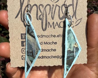 May include: A pair of triangular dangle earrings featuring a dark, abstract patterned surface with light blue edges. The earrings are attached to a brown cardboard display card printed with the text Inspired Mache and contact information.