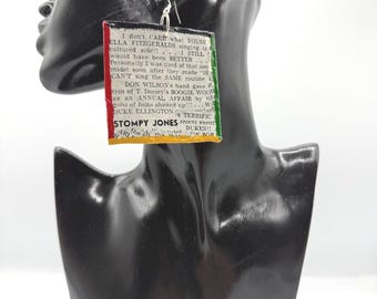 May include: A square-shaped dangle earring featuring printed vintage newspaper text. The edges are painted with red, green, and yellow stripes. The text includes mentions of Ella Fitzgerald, Duke Ellington, and Stompy Jones. It hangs from a silver-toned hook.