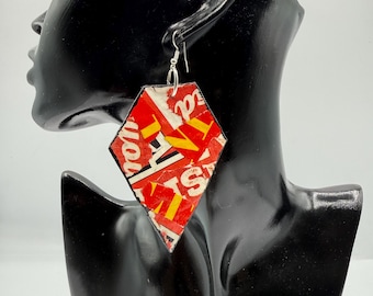 May include: A diamond-shaped dangle earring featuring a collage of red, white, and yellow printed paper fragments with visible text. The earring is suspended from a silver-toned metal hook and displays a multi-colored, layered graphic pattern.