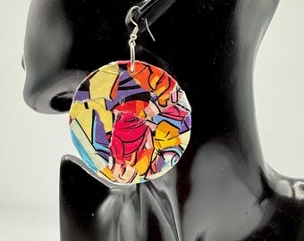 May include: A circular dangle earring featuring a vibrant collage of multicolored paper fragments in shades of red, yellow, blue, purple, and orange. The earring is suspended from a silver-toned metal hook and displays a textured, layered surface.