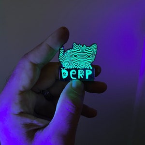 May include: A green and black enamel pin with a cartoon cat design and the word "DERP" written below it. The pin glows in the dark.