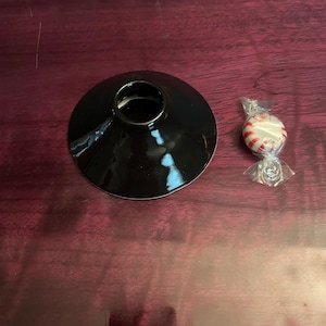 May include: A black ceramic candle holder with a wide, flat base and a small, circular opening. A wrapped peppermint candy with red and white stripes is next to the candle holder.