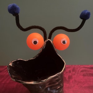 May include: A whimsical, handmade ceramic vase with a dark, textured finish. The vase is adorned with orange googly eyes, black pipe cleaner antennae, and blue pom-pom accents, creating a playful, creature-like design. The vase is suitable for home decor.