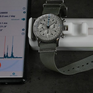 Puede incluir: Un reloj de pulsera cronógrafo plateado con una esfera blanca y agujas negras, en una correa de tela gris, se está probando en un dispositivo blanco conectado a un teléfono inteligente. La pantalla del teléfono inteligente muestra una aplicación de prueba de reloj con los resultados de la prueba, incluida la frecuencia cardíaca, el error de latido, la amplitud, el ángulo de elevación y los ruidos de latido.