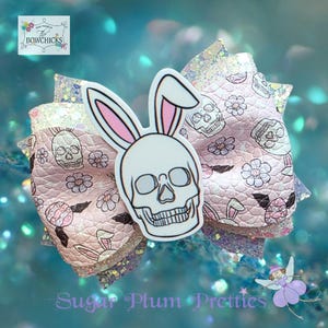 May include: A decorative hair bow featuring a white skull with bunny ears. The bow has layers of pink and glittery fabric with skull and flower patterns. The bow is a unique accessory.