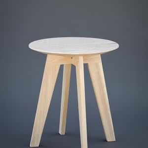 May include: A small, round wooden side table with a light-coloured finish. The table has a circular top and three angled legs. The design is minimalist and modern, suitable for various interior styles. The table's height is approximately 51 cm.
