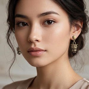 May include: A person with dark hair and brown eyes, wearing gold-coloured earrings. The earrings feature a small, detailed animal design. The person is wearing a light-coloured top, with a softly blurred background.
