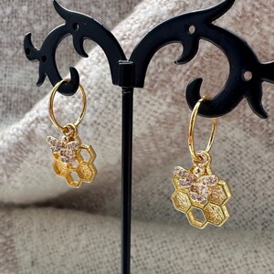 May include: A pair of gold hoop earrings with a bee and honeycomb charm. The charms are adorned with clear rhinestones.