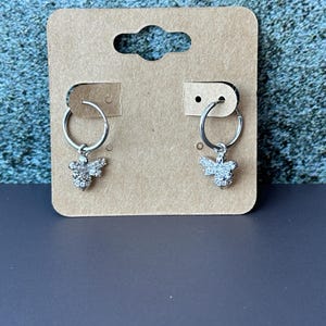 May include: A pair of silver hoop earrings with a small bee charm hanging from each hoop. The bee charms are covered in clear rhinestones.