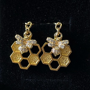 May include: Gold-toned bee earrings. Each earring features a bee charm with crystal accents, positioned above a honeycomb-shaped pendant. The earrings dangle from a small hook, set against a black background.