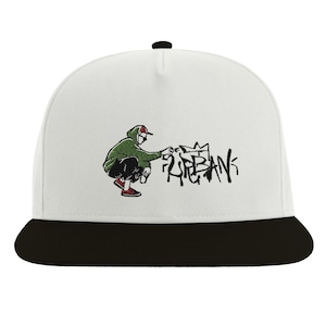Op de afbeelding: Witte en zwarte baseballpet met een geborduurd ontwerp van een persoon in een groene hoodie die graffiti spuit met de tekst "URBAN".