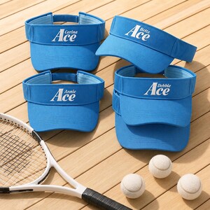 May include: Four bright blue sun visors with white embroidered text reading Ace with names Carina, Bette, Annie, and Debbie above the word. A white tennis racket and three white tennis balls are positioned near the blue athletic headwear.