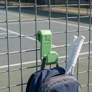 Puede incluir: Un gancho verde con la inscripción "LYDIA" está sujeto a una valla metálica, sosteniendo una mochila azul marino y dos raquetas de tenis blancas. El gancho está diseñado para colgar objetos en una valla.