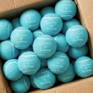 May include: A collection of bright turquoise tennis balls packed inside a cardboard box. Each ball features the white printed text "Laurent & Britney" centred on the fuzzy surface, accompanied by the standard white curved seam lines.