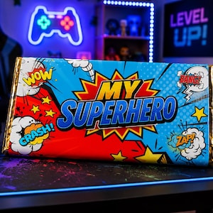 May include: A colorful candy bar with a comic book theme. The wrapper features the words "MY SUPERHERO" in bold blue letters, surrounded by action bubbles with words like "WOW" and "CRASH!" The background is red and blue.
