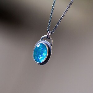 May include: A silver-toned necklace featuring a pendant with an oval, iridescent blue gemstone. The gemstone is set within a textured, silver-colored bezel and hangs from a delicate chain. The background is a soft, blurred neutral tone.