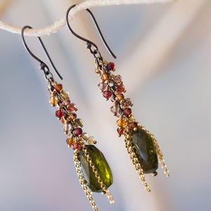 May include: A pair of dangle earrings with dark metal hooks. The earrings feature a cluster of small, colorful beads in shades of red, orange, and yellow, leading to a large, olive-green gemstone. Gold chains hang from the gemstones.