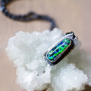 May include: A silver pendant necklace with a rectangular, iridescent stone in shades of green and blue. The pendant is set in a textured silver frame and hangs on a dark chain. The necklace rests on a white, crystalline surface.