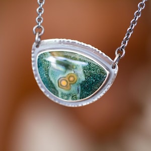 May include: A silver necklace with a pendant featuring a green and yellow stone. The stone has a unique pattern and is set in a silver bezel with a textured edge. The chain is also silver.