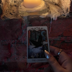 May include: A lit candle sits on a glass base, casting a warm glow against a brick wall. A tarot card, "THE CHARIOT," is held in front of the candle. The card features a black bird and a crescent moon.