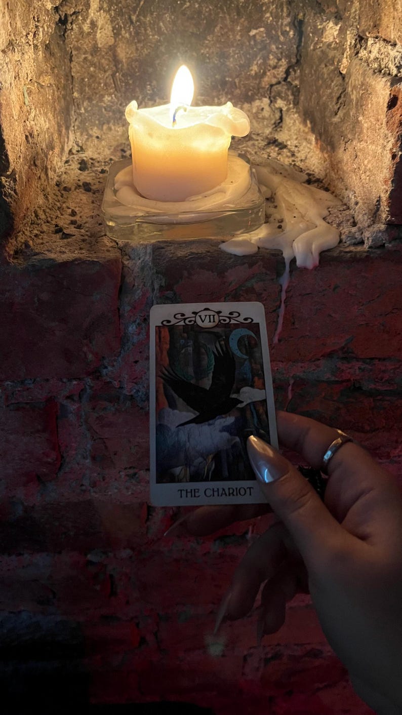 May include: A lit candle sits on a glass base, casting a warm glow against a brick wall. A tarot card, "THE CHARIOT," is held in front of the candle. The card features a black bird and a crescent moon.