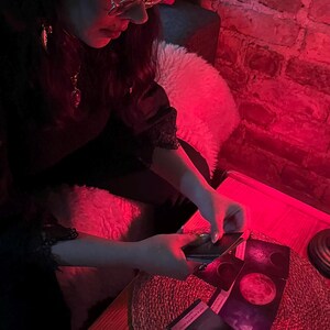 May include: A person is holding tarot cards with images of moons and celestial bodies. The scene is illuminated by red light, creating a mystical atmosphere. The cards are on a wooden table with a woven mat.