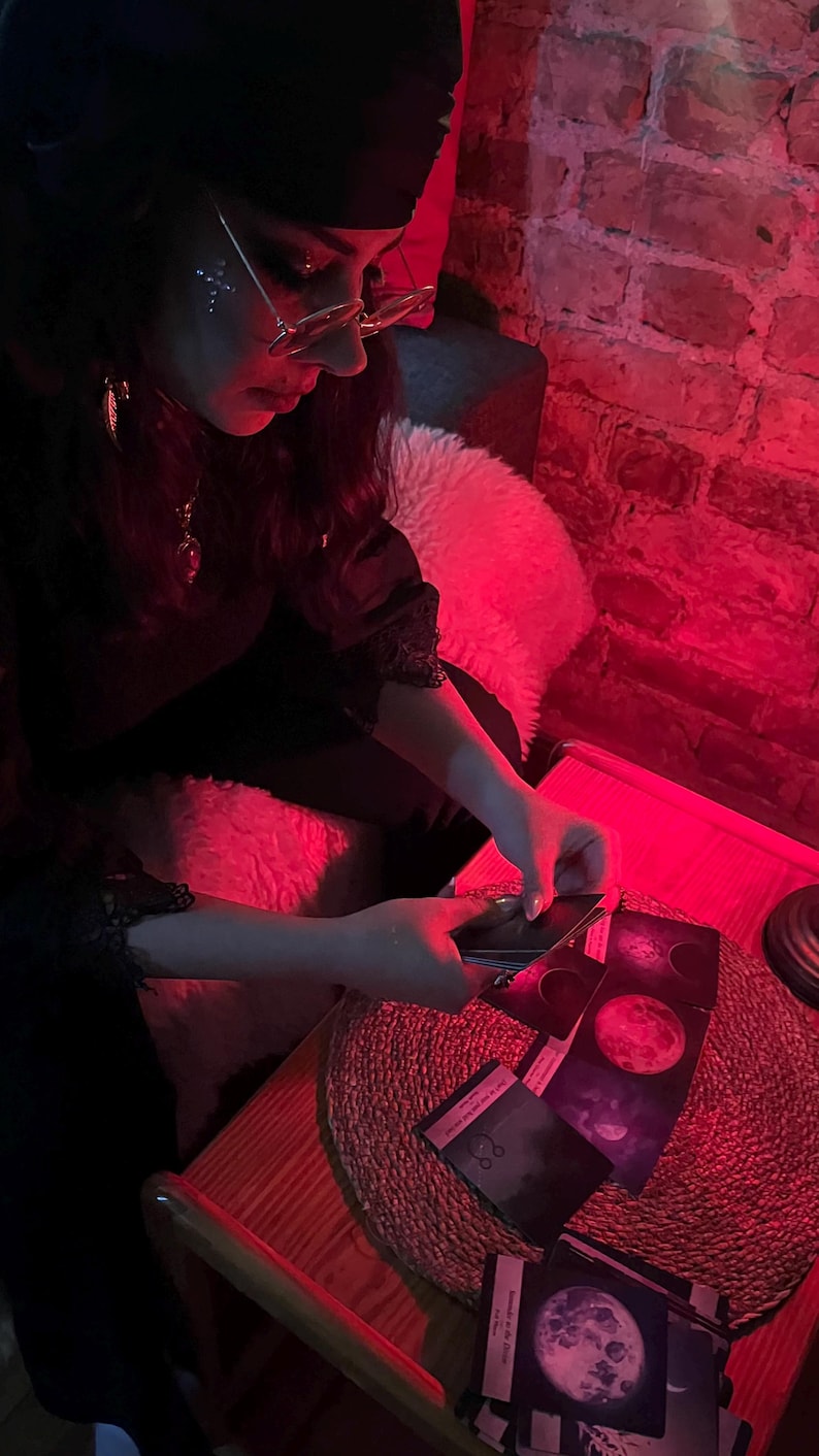 May include: A person is holding tarot cards with images of moons and celestial bodies. The scene is illuminated by red light, creating a mystical atmosphere. The cards are on a wooden table with a woven mat.