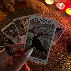 May include: A tarot card reading with the Hanged Man card prominently displayed. The card features an illustration of a figure hanging upside down. Other cards are visible, along with candles and a decorative ring.