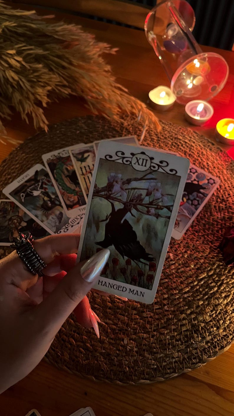 May include: A tarot card reading with the Hanged Man card prominently displayed. The card features an illustration of a figure hanging upside down. Other cards are visible, along with candles and a decorative ring.
