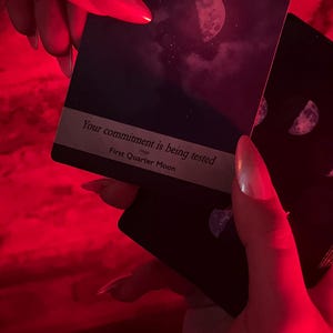 May include: Dark tarot cards with a crescent moon design and the text "Your commitment is being tested" and "First Quarter Moon". The cards are held in a red-lit environment.