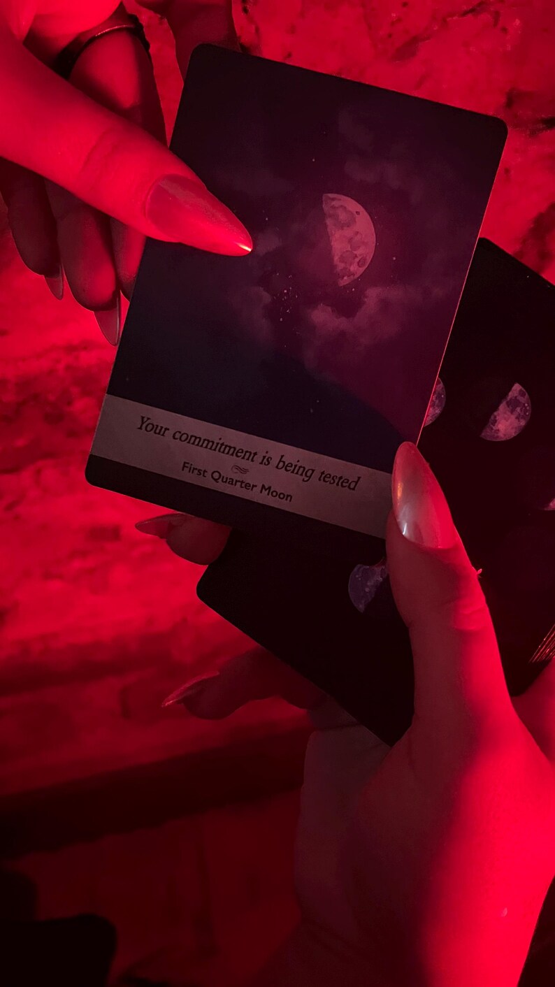 May include: Dark tarot cards with a crescent moon design and the text "Your commitment is being tested" and "First Quarter Moon". The cards are held in a red-lit environment.