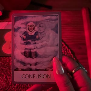 May include: A tarot card with the word "CONFUSION" at the bottom. The card depicts a person in a headscarf and dark clothing, surrounded by a cloudy, ethereal background. A second figure is visible in the background. The image is held by a hand with long, polished nails.
