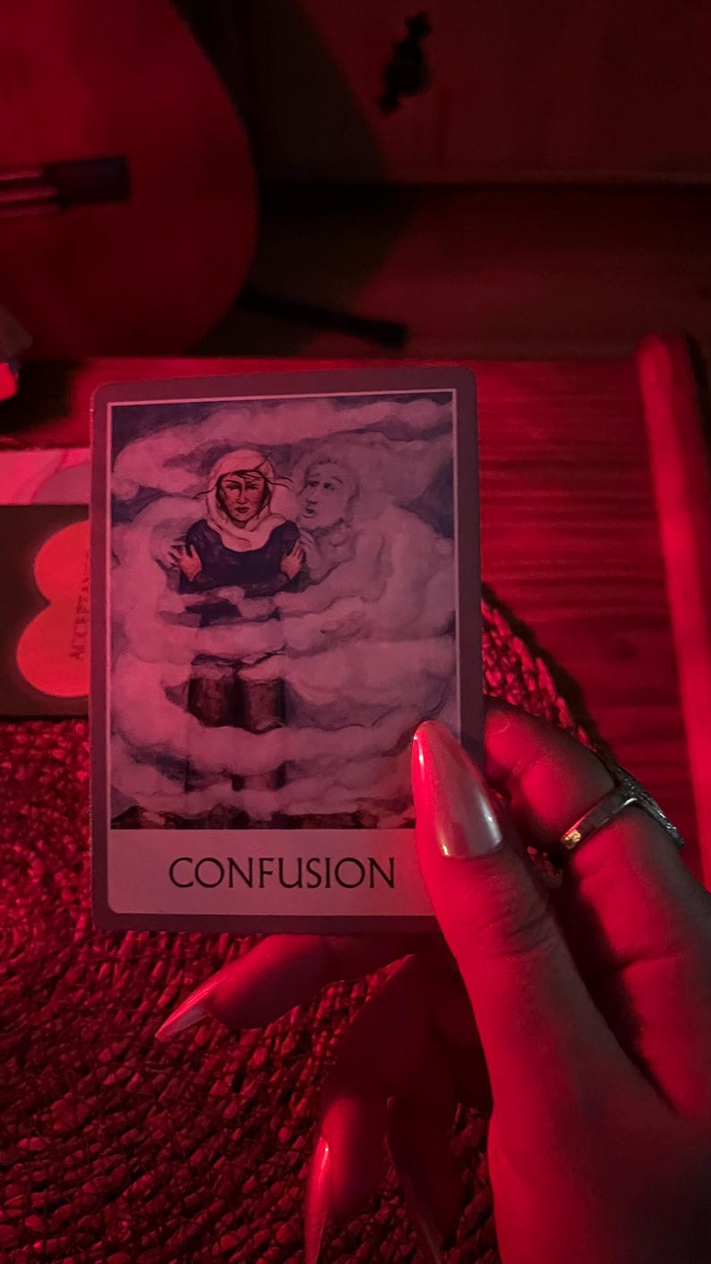 May include: A tarot card with the word "CONFUSION" at the bottom. The card depicts a person in a headscarf and dark clothing, surrounded by a cloudy, ethereal background. A second figure is visible in the background. The image is held by a hand with long, polished nails.