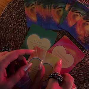 May include: A collection of oracle cards with colorful abstract designs and heart-shaped text boxes. The cards are held by a person wearing rings. The cards have words like "LOYALTY" and "DETACHMENT".