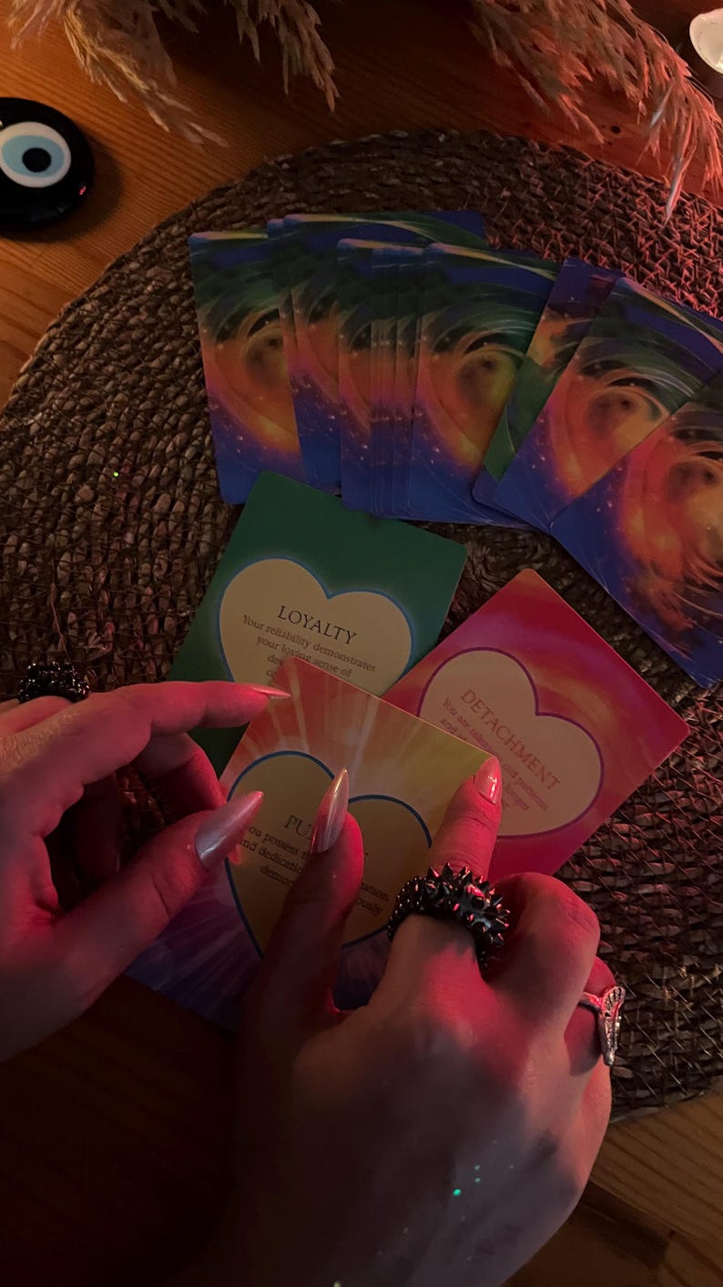 May include: A collection of oracle cards with colorful abstract designs and heart-shaped text boxes. The cards are held by a person wearing rings. The cards have words like "LOYALTY" and "DETACHMENT".