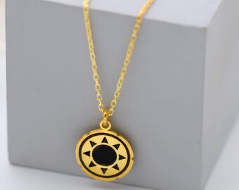 May include: A gold-colored necklace with a circular pendant featuring a black center and a sunburst design. The necklace is displayed on a gray cube, with white flowers in the background. The chain is delicate.