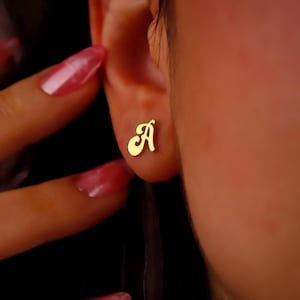 May include: Gold initial letter 'A' stud earrings. The earrings are shown in a close-up image of a person's ear.