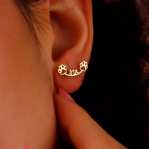May include: Gold-toned earrings with a paw print design on each side and the name "Trix" in the center.