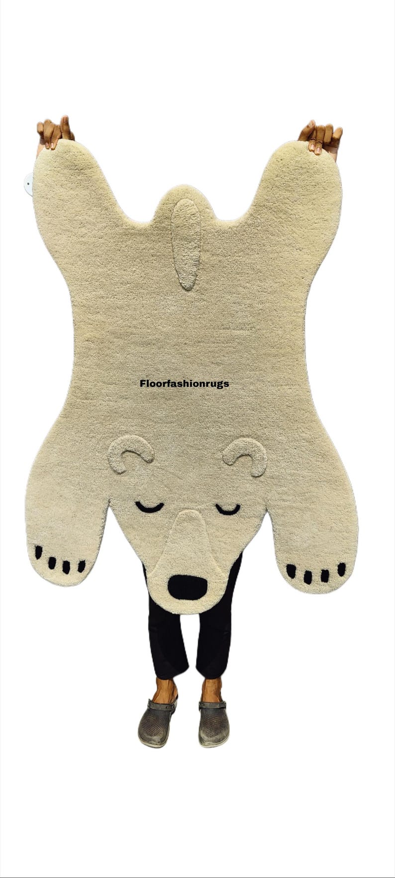 May include: A beige polar bear rug with a black nose, closed eyes, and black paw pads. The rug is held up by a person. The text "Floorfashionrugs" is visible on the rug.