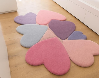 May include: A collection of heart-shaped rugs in various pastel colors, including pink, purple, and blue, arranged in a flower-like pattern. The rugs are made of a soft, textured material and are placed on a wooden floor.