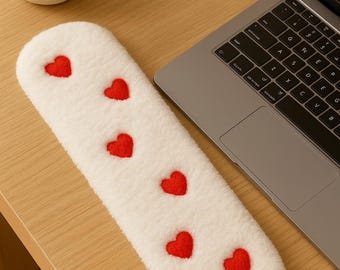 May include: A white, plush, elongated wrist rest with six red hearts embroidered on the surface. The wrist rest is designed to be placed in front of a laptop keyboard. The item is on a wooden desk.