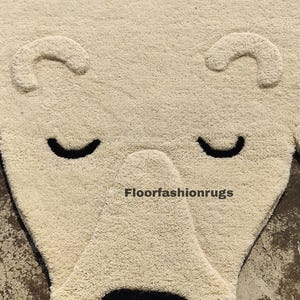 May include: A cream-colored rug shaped like a bear's face, featuring black eyes and a black open mouth. The rug has a textured surface and the text "Floorfashionrugs" is visible. This rug is suitable for a child's room or nursery.