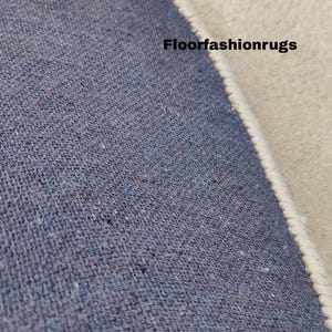 May include: Close-up of a section of a rug with a blue woven texture and a cream-colored border. The text "Floorfashionrugs" is visible on the rug's surface. The rug appears to be made of a durable material.