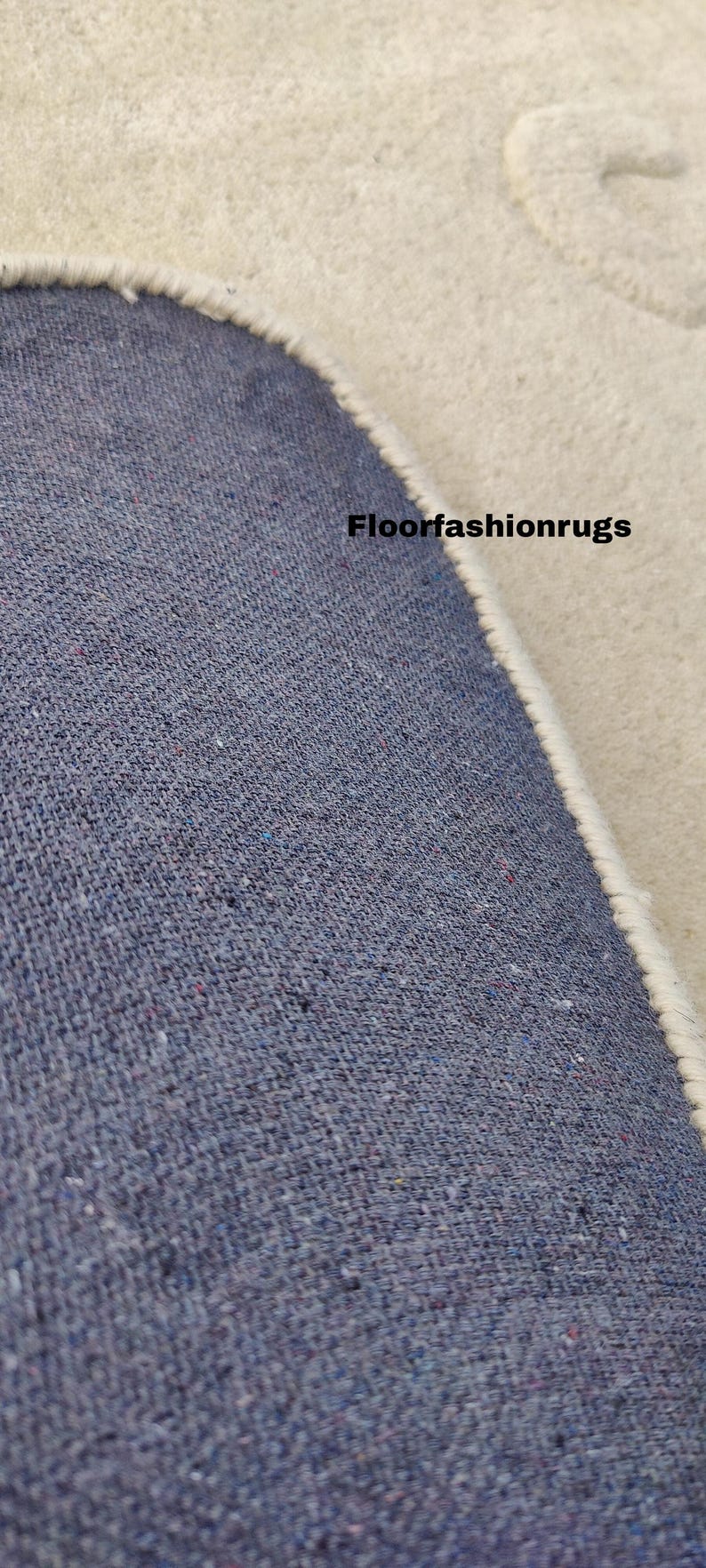 May include: Close-up of a section of a rug with a blue woven texture and a cream-colored border. The text "Floorfashionrugs" is visible on the rug's surface. The rug appears to be made of a durable material.