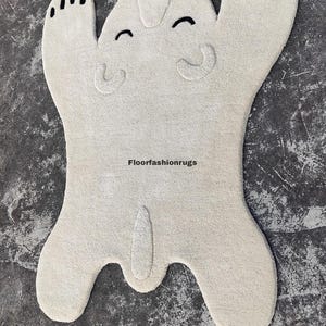 May include: A cream-colored, plush rug shaped like a cartoon bear with a smiling face and black accents. The rug has a playful design, with the text "floorfashionrugs" visible.